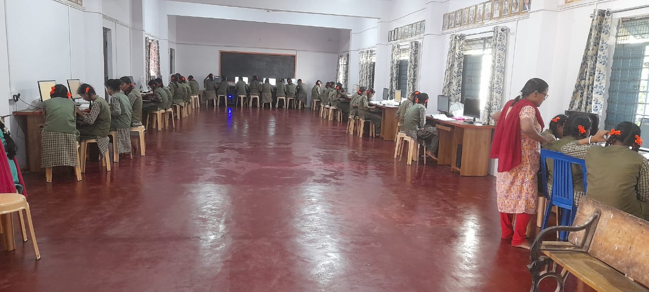 A teacher helping students at computers in a long hall