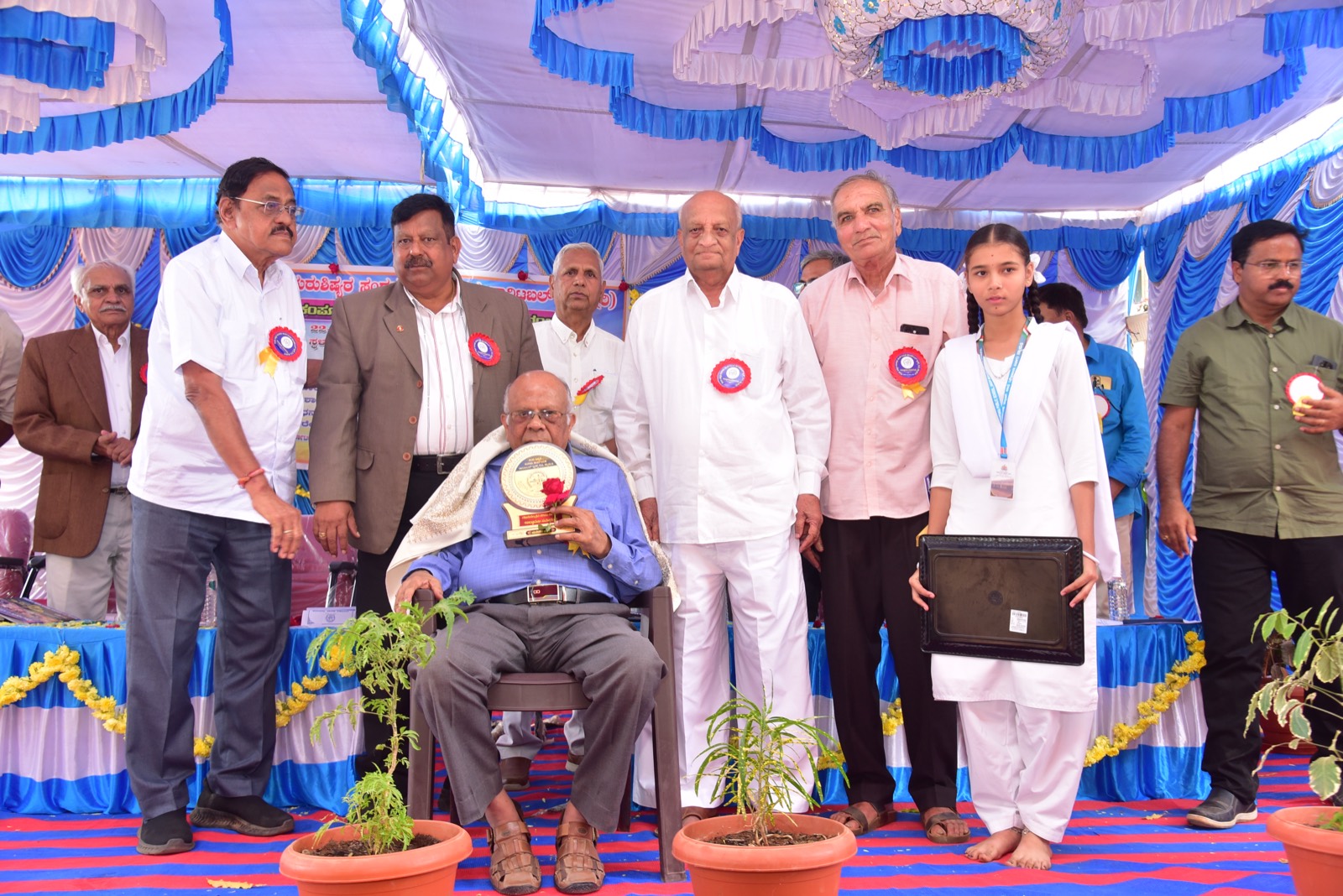 A senior guest seated on stage holding a memento, surrounded by trustees and a student